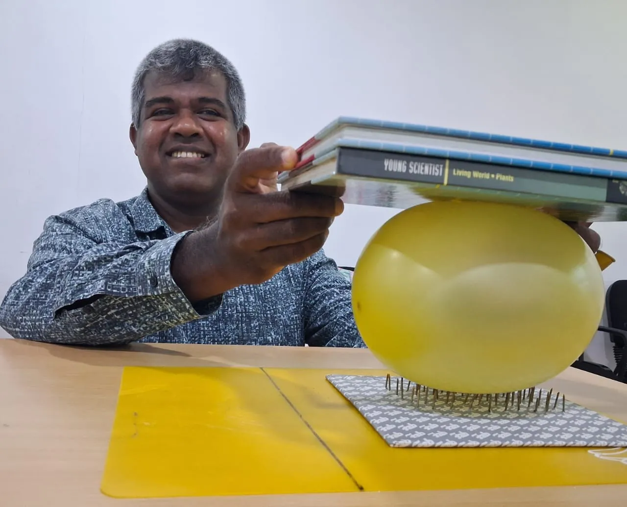 Mr. N pressing a stack of books onto a balloon resting on a bed of nails without popping it