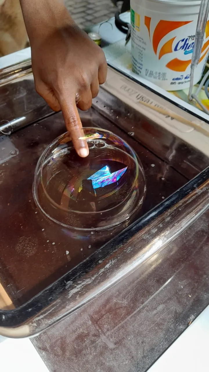A finger touching a large soap bubble dome with a tiny paper boat inside