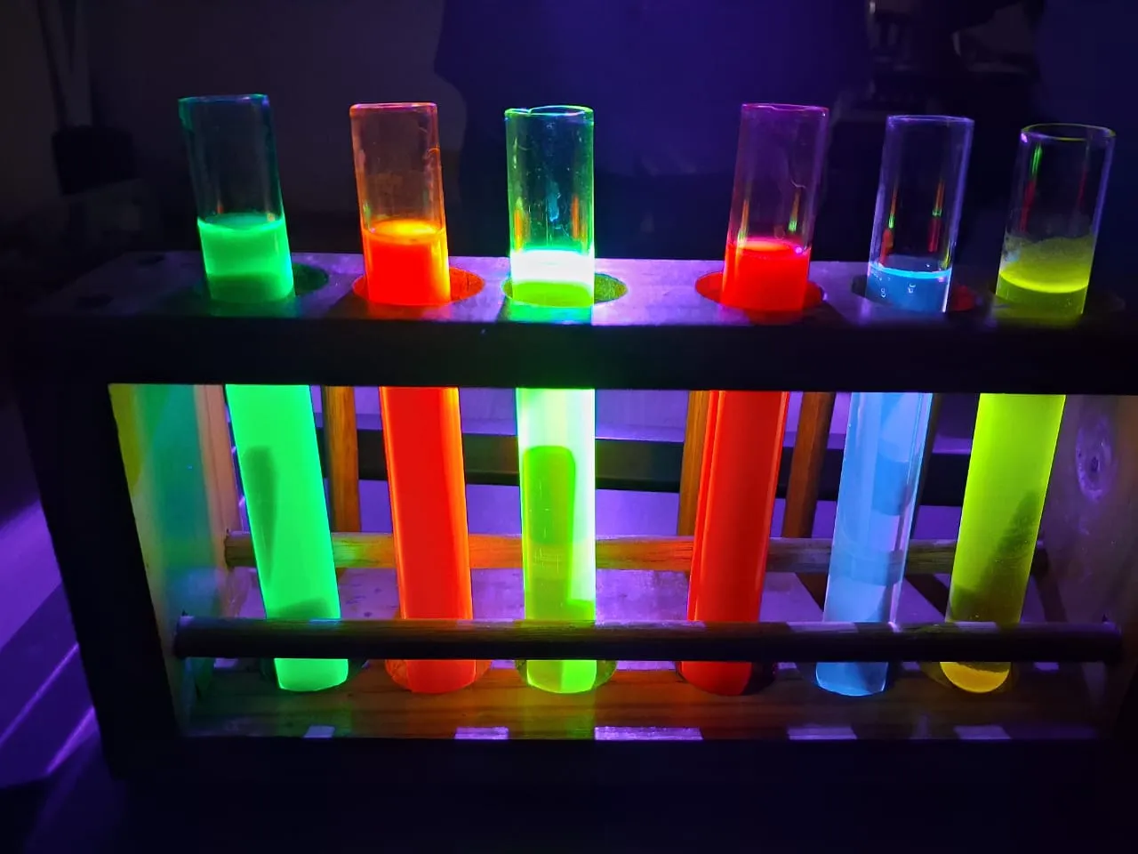 A rack of test tubes glowing neon green, orange, blue and yellow under ultraviolet light